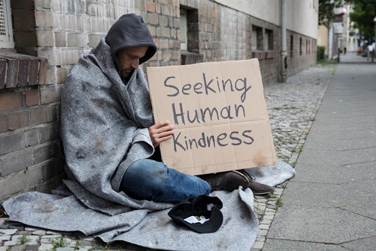 Homeless man holding placard with "Seeking Human Kindness" writing - Beddown
